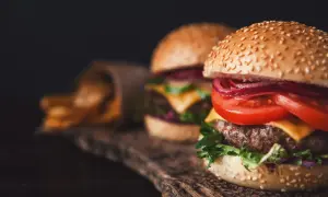 Dwa soczyste smash burgery z serem, pomidorem i czerwoną cebulą, obok papierowego rożka z frytkami.