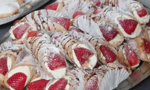 Taca pełna pysznych cannoli z kremem i truskawkami, posypana cukrem pudrem i polana czekoladą.
