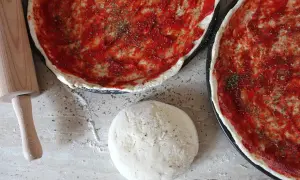 Jak zrobić ciasto na pizzę? Na blacie leży kulka ciasta, wałek i dwie blachy z sosem pomidorowym i ziołami.