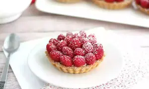 Pyszne tartaletki z malinami, posypane cukrem pudrem, na białym talerzyku. Obok łyżeczka i serwetka w kwiatki.