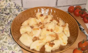 Pyszne kopytka ruskie polane skwarkami z cebulki, podane na brązowym talerzu, obok gałązka pomidorków koktajlowych.