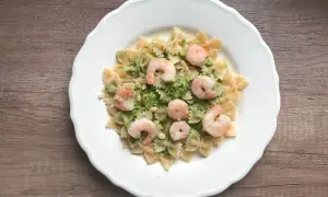 Makaron farfalle z zielonym pesto, krewetkami i orzeszkami pinii na białym talerzu.
