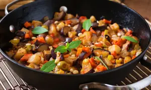 Pyszna caponata z bakłażana z oliwkami i orzeszkami pinii, podana w czarnej patelni, ozdobiona listkami bazylii.