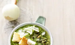 Aksamitny krem z czosnku niedźwiedziego, udekorowany grzankami, pestkami dyni i parmezanem. Obok leży cebula.