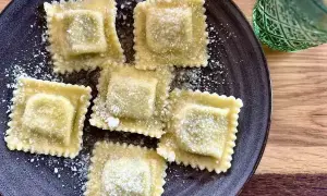 Pięć kwadratowych ravioli z ricottą, posypanych parmezanem, na ciemnym talerzu. Obok zielony kieliszek.