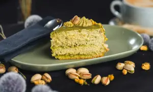 Pyszny kawałek tortu pistacjowego, udekorowany kremem i orzechami. Obok filiżanka kawy i łuskane pistacje.