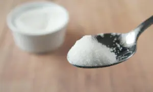 Łyżeczka białego, drobnego cukru, jakby cukier bez cukru. W tle biała miseczka z tym samym produktem.