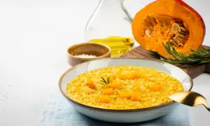 Kremowe risotto dyniowe w misce, z kawałkami dyni i rozmarynem. Obok połówka dyni, oliwa i sól.