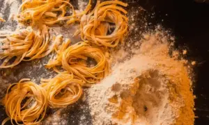 Świeże, domowe tagliatelle uformowane w kółka, leżą obok kopczyka mąki na ciemnym blacie.