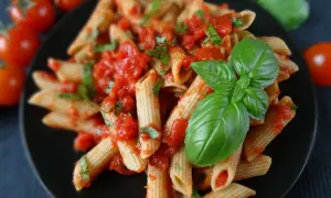 Penne z sosem pomidorowym i świeżą bazylią, ozdobione listkami bazylii. Obok pomidorki koktajlowe.