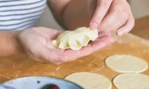 Ręce formują pieroga według przepisu. Na blacie leżą okrągłe placki ciasta i miska z wiśniami.