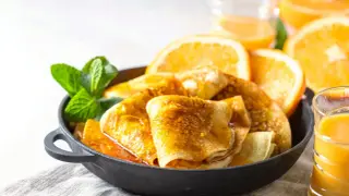 Złociste crepes suzette w sosie pomarańczowym, udekorowane miętą i plastrami pomarańczy. Obok szklanka soku pomarańczowego.