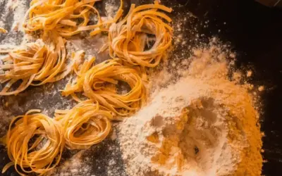 Świeże, domowe tagliatelle uformowane w kółka, leżą obok kopczyka mąki na ciemnym blacie.