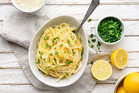 Pyszny makaron cytrynowy posypany parmezanem i świeżą natką pietruszki, obok połówki cytryny.