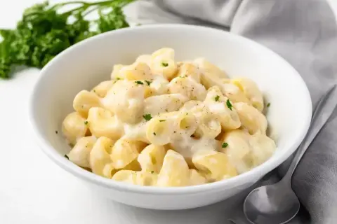Miseczka kremowego mac and cheese, posypana natką pietruszki, obok łyżka i zielona gałązka.