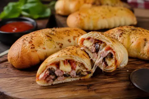 Złociste pierogi pizza z ciągnącym serem, szynką i pieczarkami, podane na desce z sosem pomidorowym.
