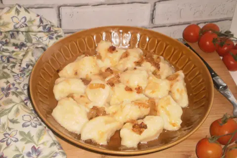 Pyszne kopytka ruskie polane skwarkami z cebulki, podane na brązowym talerzu, obok gałązka pomidorków koktajlowych.