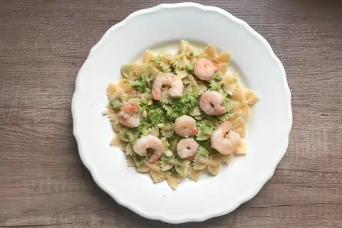 Makaron farfalle z zielonym pesto, krewetkami i orzeszkami pinii na białym talerzu.