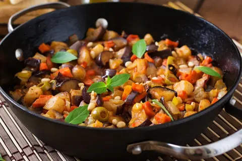 Pyszna caponata z bakłażana z oliwkami i orzeszkami pinii, podana w czarnej patelni, ozdobiona listkami bazylii.