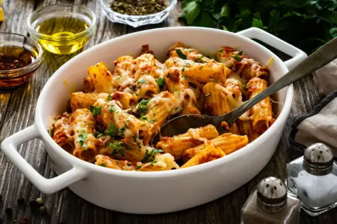 Pyszny obiad z mięsa mielonego: makaron rigatoni zapieczony z sosem bolońskim i ciągnącym serem, posypany świeżą natką pietruszki.