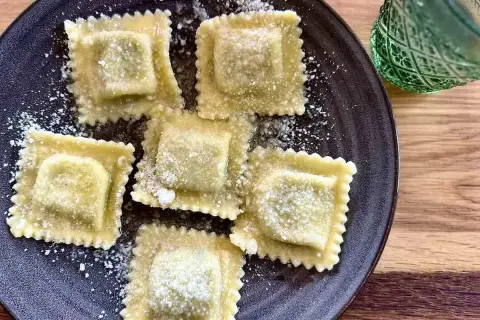 Pięć kwadratowych ravioli z ricottą, posypanych parmezanem, na ciemnym talerzu. Obok zielony kieliszek.
