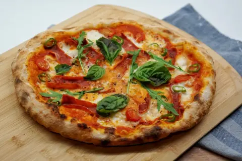 Pyszna pizza z rukolą, pomidorkami, papryczką i wędliną. Idealne dodatki do pizzy na drewnianej desce.