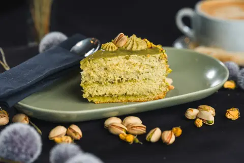 Pyszny kawałek tortu pistacjowego, udekorowany kremem i orzechami. Obok filiżanka kawy i łuskane pistacje.