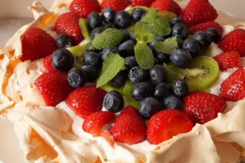 Pyszny tort bezowy z owocami: truskawki, borówki, kiwi i listki mięty na kremie.