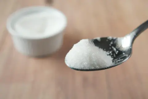 Łyżeczka białego, drobnego cukru, jakby cukier bez cukru. W tle biała miseczka z tym samym produktem.