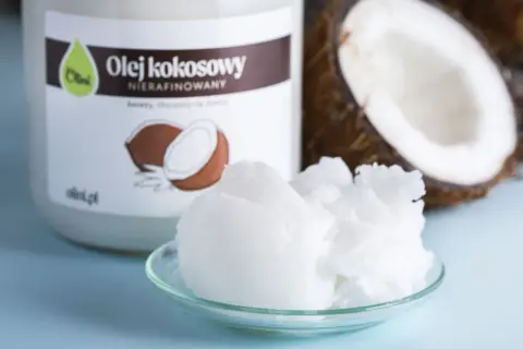 Olej kokosowy nierafinowany na talerzyku, idealny jako jaki olej do smażenia. Obok połówka kokosa i słoik z etykietą.