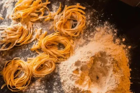 Świeże, domowe tagliatelle uformowane w kółka, leżą obok kopczyka mąki na ciemnym blacie.