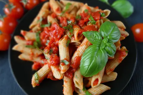 Penne z sosem pomidorowym i świeżą bazylią, ozdobione listkami bazylii. Obok pomidorki koktajlowe.