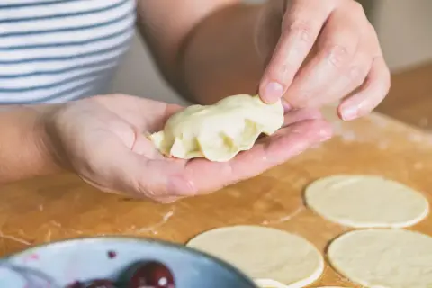 Ręce formują pieroga według przepisu. Na blacie leżą okrągłe placki ciasta i miska z wiśniami.