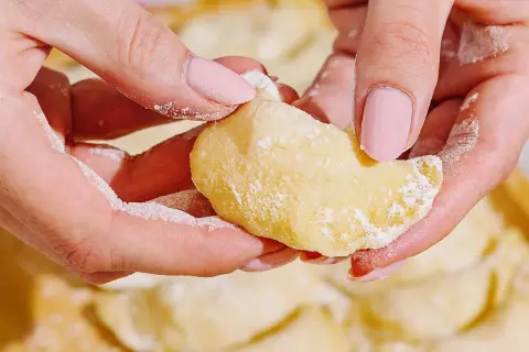 Dłonie z różowymi paznokciami formują ciasto do pierogów, obsypane mąką. W tle widać gotowe pierogi.