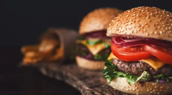 Dwa soczyste smash burgery z serem, pomidorem i czerwoną cebulą, obok papierowego rożka z frytkami.