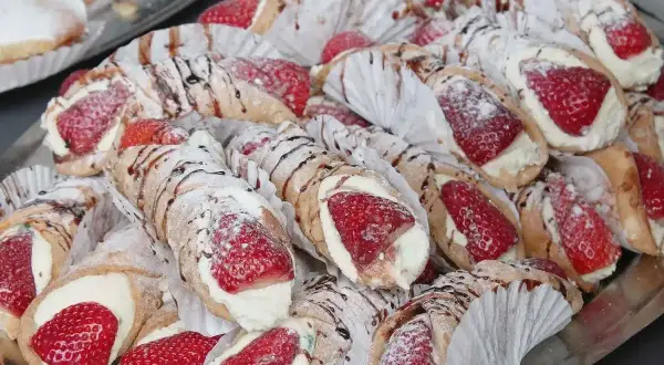 Taca pełna pysznych cannoli z kremem i truskawkami, posypana cukrem pudrem i polana czekoladą.