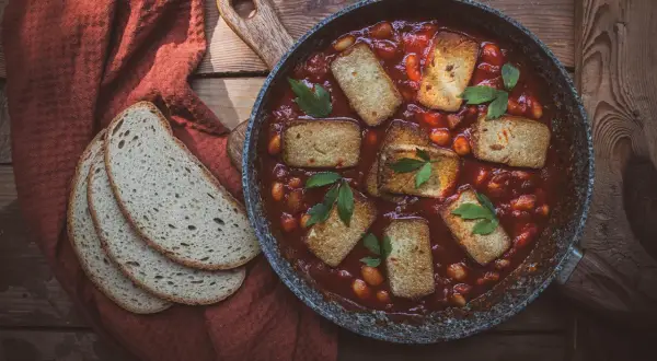 Aksamitne tofu w sosie pomidorowym z fasolką, podane w patelni, obok kromek chleba na rustykalnym stole.