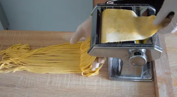 Ręce w rękawiczkach formują prosta pasta fresca na maszynce do makaronu. Świeży, żółty makaron opada na drewniany blat.