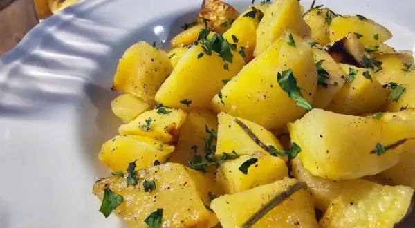 Pieczone ziemniaki pokrojone w ćwiartki, posypane natką pietruszki i pieprzem, podane na białym talerzu.
