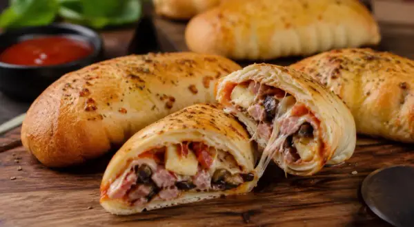 Złociste pierogi pizza z ciągnącym serem, szynką i pieczarkami, podane na desce z sosem pomidorowym.