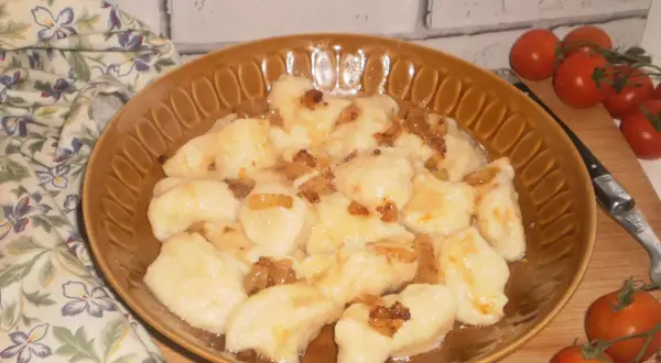 Pyszne kopytka ruskie polane skwarkami z cebulki, podane na brązowym talerzu, obok gałązka pomidorków koktajlowych.