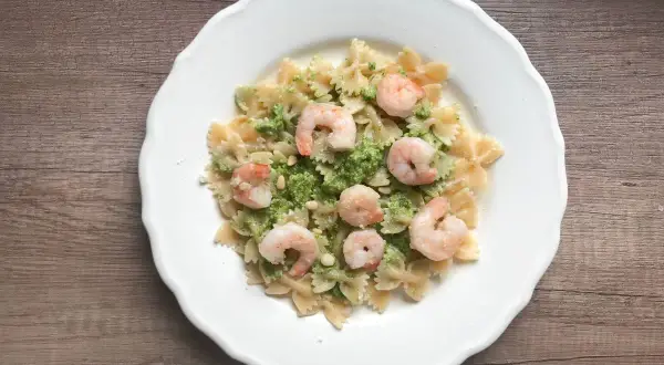 Makaron farfalle z zielonym pesto, krewetkami i orzeszkami pinii na białym talerzu.