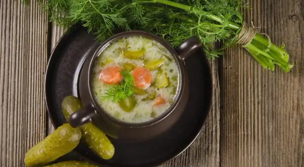 Aromaticzna zupa ogórkowa na żeberkach, podana w brązowej miseczce, ozdobiona koperkiem. Obok leżą chrupiące ogórki kiszone i pęczek świeżego koperku.