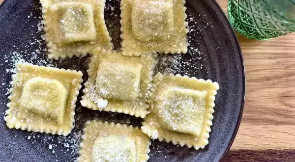 Pięć kwadratowych ravioli z ricottą, posypanych parmezanem, na ciemnym talerzu. Obok zielony kieliszek.