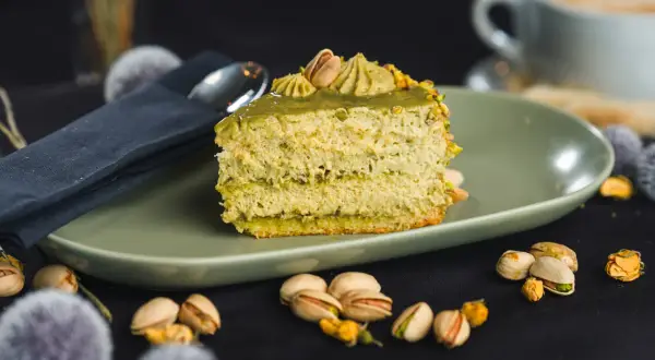 Pyszny kawałek tortu pistacjowego, udekorowany kremem i orzechami. Obok filiżanka kawy i łuskane pistacje.