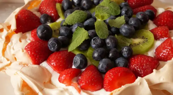 Pyszny tort bezowy z owocami: truskawki, borówki, kiwi i listki mięty na kremie.