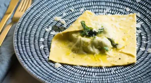 Duży raviolo con uovo, polany masłem i posypany parmezanem, na talerzu z czarno-białym wzorem.
