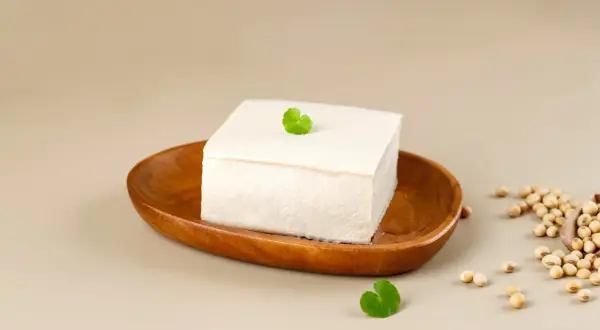 Kostka białego tofu na drewnianej desce, obok nasiona soi. Różne rodzaje tofu są wszechstronne w kuchni.