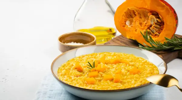 Kremowe risotto dyniowe w misce, z kawałkami dyni i rozmarynem. Obok połówka dyni, oliwa i sól.