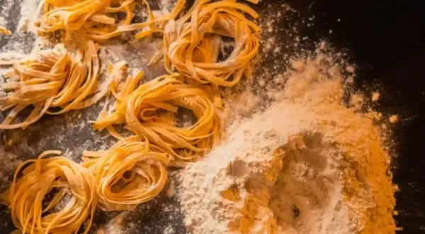 Świeże, domowe tagliatelle uformowane w kółka, leżą obok kopczyka mąki na ciemnym blacie.